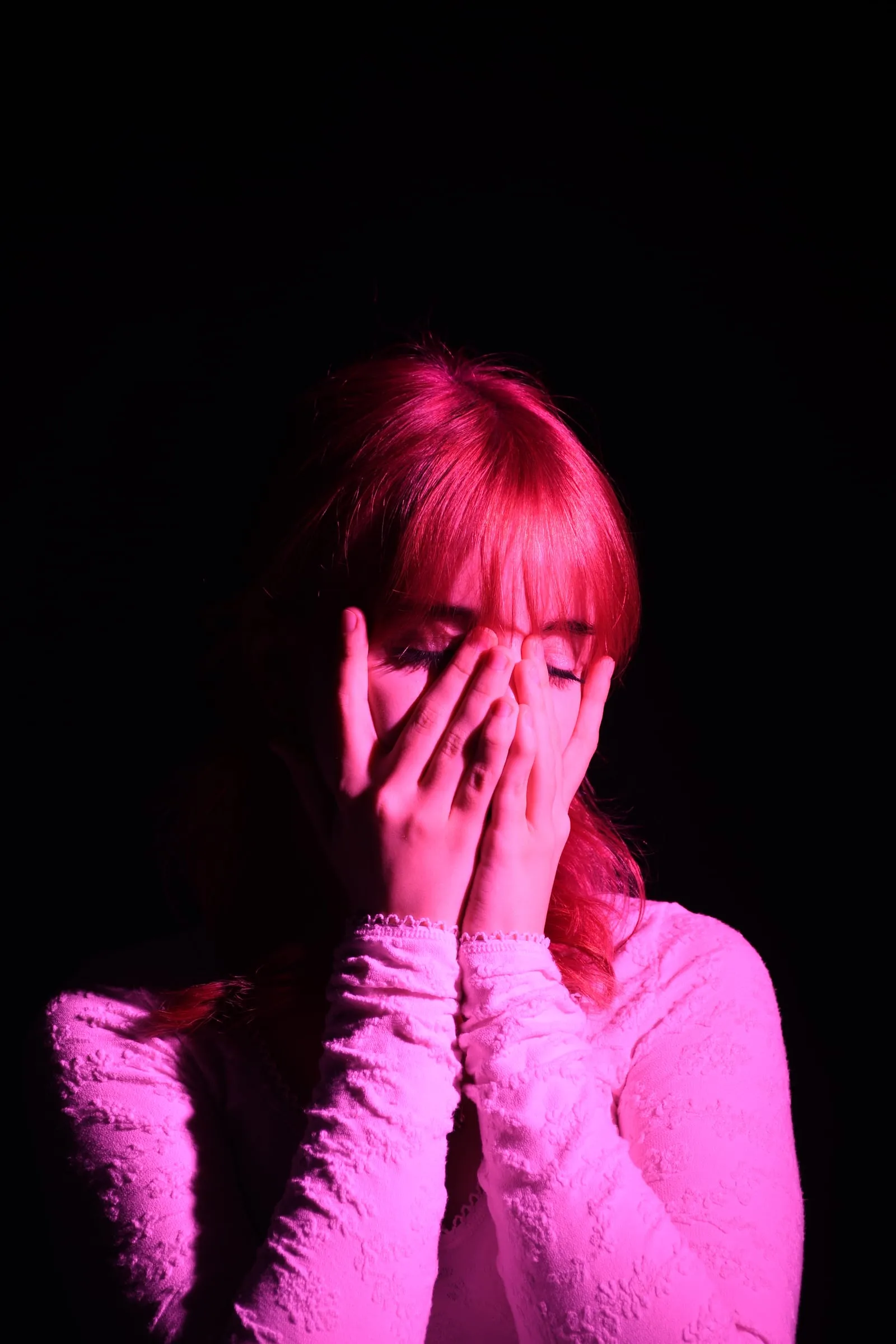 A woman hides her face in her hands under magenta light.